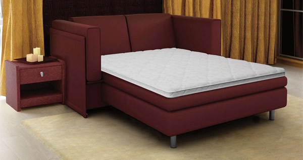 Dark, low-key interior photograph of a bed with a deep red upholstered frame, white quilted top, and warm candlelit nightstand